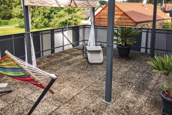 The rooftop terrace with old concrete paving offers a spacious area to relax. The dirty surface of the concrete slabs shows clear signs of wear. The sunshade provides pleasant shade on hot days. Plant pots with palms and a hammock create a welcoming atmosphere. The metal and glass railing completes the modern, open design of the terrace.