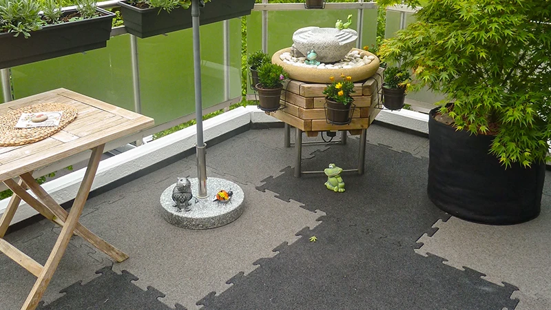 This photo shows an inviting balcony laid with two-tone WARCO puzzle tiles. A small fountain, a wooden shelf, and several planters create a cosy atmosphere. The flooring offers high comfort with its elastic, slip-resistant surface and is also weatherproof. Thanks to precise interlocking, it allows for accent colours that give the outdoor area a modern flair.