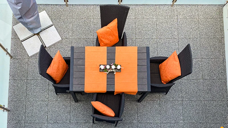 This image shows a spacious balcony featuring grey WARCO floor tiles. A large dining table sits at the centre, surrounded by black rattan chairs accented with orange cushions. The elastic, slip-resistant flooring offers comfort and secure footing. In addition, it is low-maintenance and weatherproof, giving the outdoor space a modern feel that encourages relaxation.