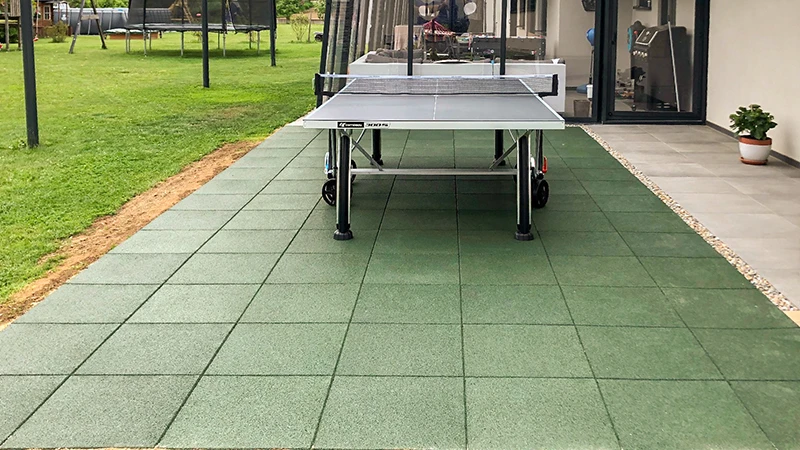 In a lush garden in front of a luxurious house, a terrace of green WARCO tiles made of PU-bound rubber granulate accommodates a ping-pong table. Nearby, a trampoline and other play equipment invite active recreation for all ages. The robust, slip-resistant surface ensures comfort and safety, even in changing weather. This durable flooring offers easy maintenance and enhances the outdoor experience, combining functionality with a fresh, inviting design.