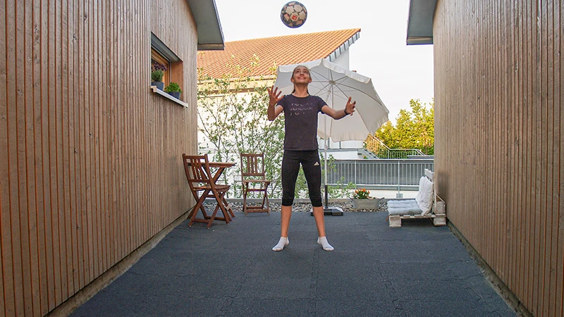 A girl is playing ball on a terrace between two wooden-clad building walls. The flooring consists of black rubber granulate terrace tiles, providing a robust and resilient surface suitable for sports activities. In the background, a sunshade and a small wooden seating area create an inviting atmosphere.