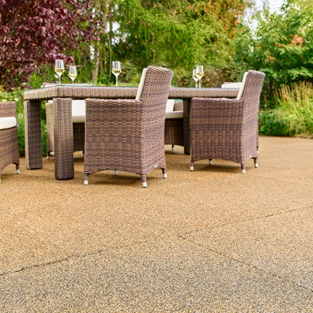 Sunlit garden patio featuring beige-brown WARCO tiles made from PU-bound rubber granules. The slip-resistant, water-permeable surface ensures a comfortable walking experience and reduces noise. A sturdy garden table with woven chairs and filled wine glasses invites relaxation. Vibrant shrubs and colorful flowers create a natural setting. This elastic flooring is frost-resistant, retains its shape, and provides long-lasting durability. Ideal for year-round use, it complements any outdoor décor.