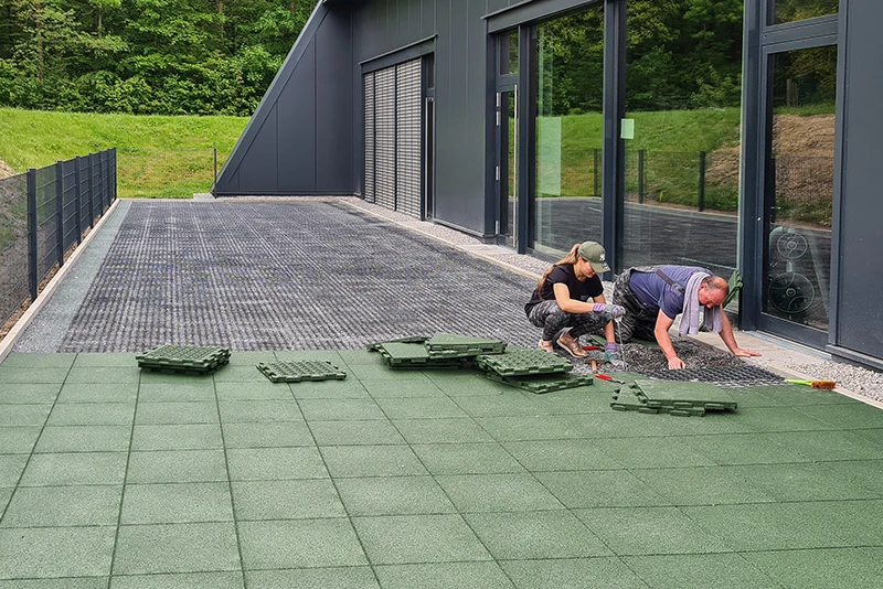 Against a dark, modern façade, a woman and a man are installing green WARCO patio tiles made from PU-bound rubber granulate. The tiles fit together seamlessly to create a slip-resistant, weatherproof and durable surface for the terrace. This sturdy material provides comfort and protection in all weathers. The straightforward installation process enables personalised layouts, enhancing any terrace with a high-quality, contemporary finish.
