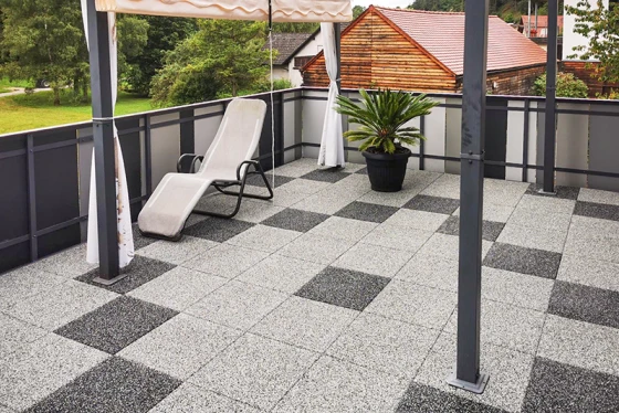 The renovated rooftop terrace features granite-look paving tiles arranged in a geometric pattern of light and dark grey squares, creating a modern and stylish atmosphere. The robust rubber tile flooring provides slip resistance and comfort. A sunshade offers pleasant shade on hot days. A planter with a palm and a lounger complete the inviting terrace design. The metal and glass railing gives the rooftop terrace an open and airy appearance.