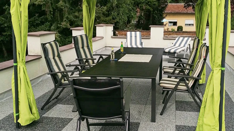 On a rooftop terrace, a flooring made of WARCO terrace tiles features a light and dark grey marbled look reminiscent of granite. A canopy shades the seating area on the elastic, weather-resistant surface. The slip-resistant, shock-absorbing tiles can be installed without permanent adhesion and help insulate footstep noise. The result is a low-maintenance space perfect for relaxing outdoors.