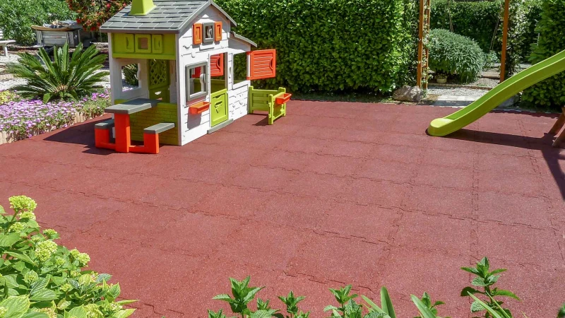 Playground in the garden In the middle of the garden, surrounded by shrubs, there is a play area made of brick-red WARCO playground flooring. A colorful playhouse and a children's slide are available for the children to play on.