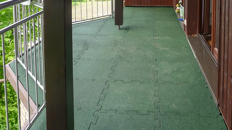 This photo shows a long balcony fitted with green WARCO flooring tiles in puzzle form. The slip-resistant and elastic surface provides safety and comfort, even in wet conditions. Thanks to the precise interlocking design, the result is a durable, low-maintenance solution that protects the subfloor. This creates a pleasant outdoor area, perfect for relaxing and enjoying the fresh air.