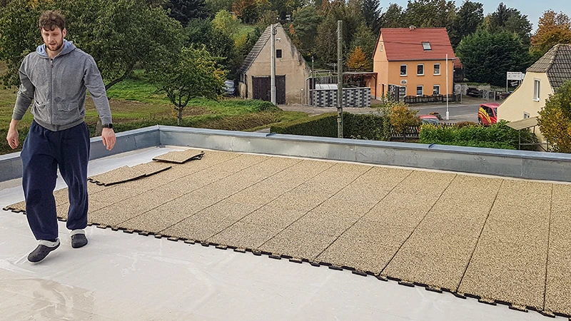 In this photo, a person is installing beige WARCO puzzle tiles on a generously sized balcony. The precise interlocking system allows for quick and easy assembly of the flooring. The slip-resistant, weatherproof surface protects the subfloor and provides a walkable area for multiple purposes. This results in a safe, comfortable, and low-maintenance outdoor space, perfect for relaxation. Additionally, its insulating properties help reduce footstep noise.
