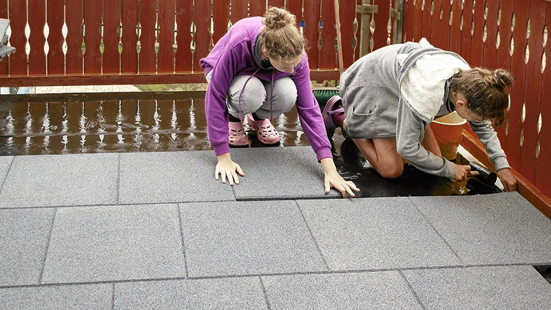 In this photo, two individuals are installing grey WARCO flooring tiles on a wet wooden deck. Thanks to their precise fit, the tiles can be laid quickly and easily. The slip-resistant, water-permeable surface is ideal for outdoor areas. These durable tiles protect the underlying structure and ensure a safe, comfortable walking experience. The result is a sturdy, low-maintenance terrace in no time.