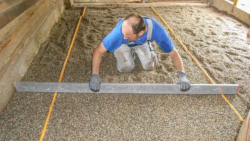Installation of a new floor in a horse stable: A man uses a screed to level a gravel sub-base. The gravel serves as a bedding layer for the installation of a rubber granulate stable floor. The area is bordered by timber or concrete walls. The work is done with care and precision – typical for the proper preparation of a horse-friendly stable floor.