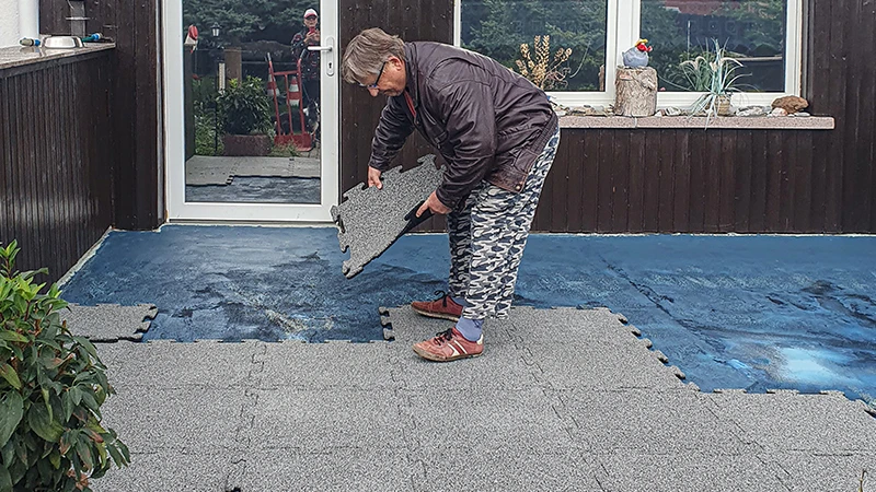 A terrace is being renovated. An elderly woman is laying the next 18 mm thick terrace tile with interlocking edges onto the already partially covered terrace. A sealing membrane serves as a substrate for the new rubber tile flooring. The precise interlocking of the tiles allows for easy and stable installation.
