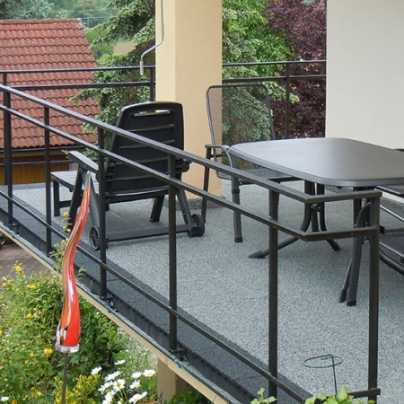 This photo shows a large balcony covered with grey WARCO flooring tiles. A black metal railing encloses the space, providing safety. A generously sized table and chairs invite social gatherings. The elastic, slip-resistant surface is weatherproof and easy to maintain, ensuring lasting protection for this outdoor area while offering a comfortable spot to relax.
